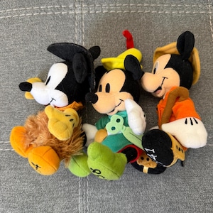 K&ouml;nnte beinhalten: Drei Pl&uuml;schtiere, die Mickey Mouse in verschiedenen Outfits darstellen. Die linke Figur tr&auml;gt einen schwarzen Hut und einen braunen, fellartigen K&ouml;rper. Die mittlere tr&auml;gt einen gelben Hut und ein gr&uuml;nes Outfit. Die rechte tr&auml;gt einen beigen Hut und eine orangefarbene Jacke.