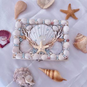Puede incluir: Una caja rectangular decorada con conchas marinas blancas, una estrella de mar central y perlas azul claro. La tapa presenta una concha grande en forma de abanico rodeada de conchas pequeñas y cuentas. Otras conchas rodean la caja.