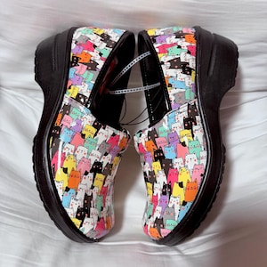May include: Pair of colorful clogs with a black sole. The clogs are covered in a pattern of various cartoon cats in shades of pink, yellow, blue, green, and black. The shoes have a slight heel and a rounded toe.
