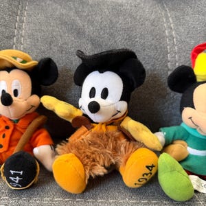 K&ouml;nnte beinhalten: Drei Mickey Mouse Pl&uuml;schfiguren. Die linke Figur tr&auml;gt einen orangefarbenen Mantel mit der Zahl 941 auf dem Fu&szlig;. Die mittlere Figur tr&auml;gt einen schwarzen Hut und eine braune fellartige Hose mit der Zahl 1934 auf dem Fu&szlig;.