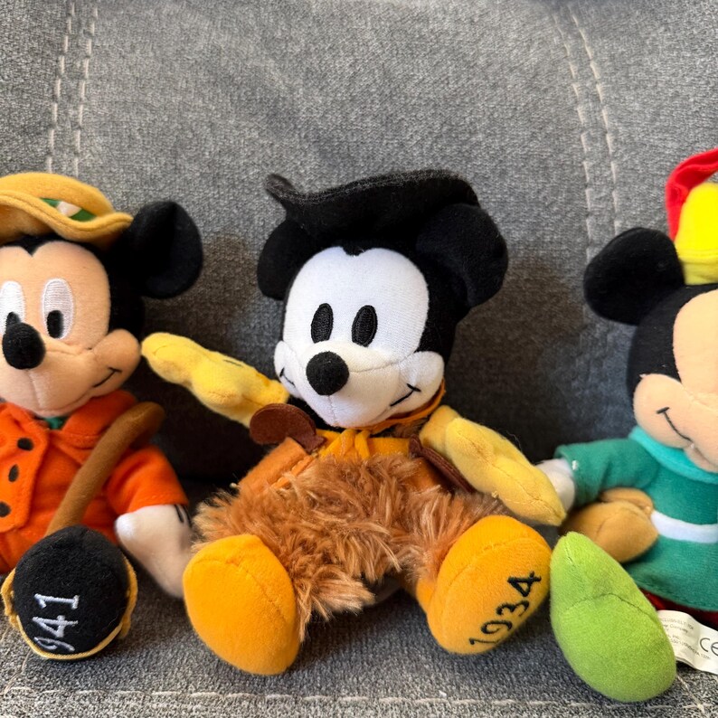 K&ouml;nnte beinhalten: Drei Mickey Mouse Pl&uuml;schfiguren. Die linke Figur tr&auml;gt einen orangefarbenen Mantel mit der Zahl 941 auf dem Fu&szlig;. Die mittlere Figur tr&auml;gt einen schwarzen Hut und eine braune fellartige Hose mit der Zahl 1934 auf dem Fu&szlig;.