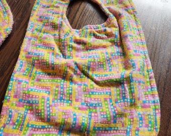 May include: A colorful baby bib with a geometric pattern in yellow, pink, blue, and green. The bib has a rounded shape with a neck opening and a yellow snap closure. The fabric appears soft and absorbent.