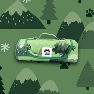 Op de afbeelding: Een opgerolde groene dierenmat met een handvat, met een berg- en boompatroon. De mat heeft een groene rand en zilveren drukkers. Het Paw Hub-logo is zichtbaar op de voorkant. De achtergrond is een herhalend patroon van bergen, bomen en pootafdrukken.