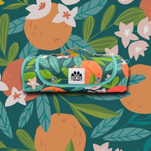 Op de afbeelding: Een opgerolde dierenmat met een turkooizen rand en handvat, met een levendig patroon van sinaasappels, bladeren en bloemen. De mat heeft een flap die met drukknopen is vastgezet en een "PAW HUB"-logo. De achtergrond is een bijpassend bloemmotief.