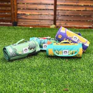 May include: Four rolled-up pet blankets in various colors, including green, teal, yellow, and purple, are displayed on a green lawn. Each blanket features a handle and a paw print logo. The blankets have different patterns, including trees, floral designs, and text.