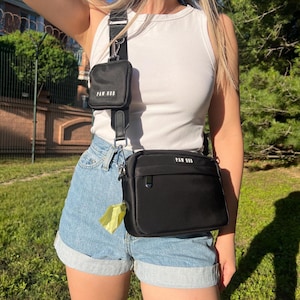 Peut inclure: Sac bandoulière noir avec une petite pochette assortie, toutes deux avec le texte "PAW HUB". Le sac a une bandoulière noire réglable et un petit distributeur de sacs jaune attaché. La personne porte un débardeur blanc et un short en jean.