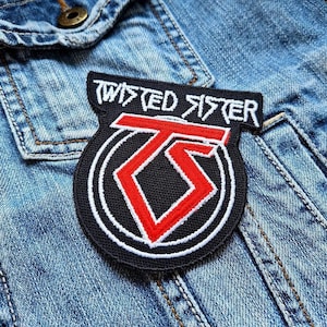 Puede incluir: Parche bordado en negro y rojo con el texto "TWISTED SISTER" en blanco. El parche presenta un logotipo "TS" rojo dentro de un círculo negro con borde blanco. El parche está cosido en una chaqueta vaquera.