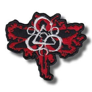 May include: Embroidered patch featuring a stylized design in red, black, and white. The patch has a unique shape with a central emblem composed of interconnected shapes and lines. The design is set against a black background with red accents.