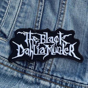 Könnte beinhalten: Schwarzer, gestickter Aufnäher mit dem weißen Schriftzug "The Black Dahlia Murder" in stilisierter Schrift. Der rechteckige Aufnäher hat einen schwarzen Rand und ist auf eine Jeansjacke genäht.