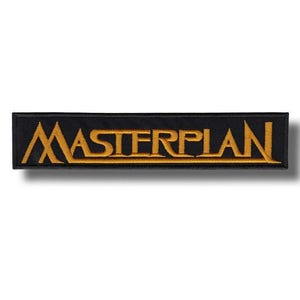 May include: A rectangular black patch with the word "MASTERPLAN" embroidered in gold thread. The patch has a simple, clean design, with the text centered on the black background. The patch is likely for clothing or accessories.