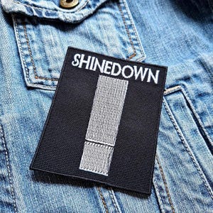 以下が含まれることがあります： 白文字で「SHINEDOWN」と書かれた黒い長方形のパッチ。白い縦のデザインがその下にあります。パッチは明るい青色のデニムジャケットに縫い付けられています。
