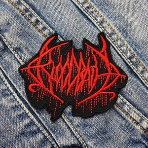 May include: A black patch with red embroidered text. The text is stylized and appears to be a band logo or similar design. The patch is on a denim background.
