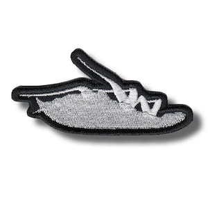 Puede incluir: Parche bordado con borde negro e interior plateado texturizado. Un elemento negro se extiende hacia arriba. El parche está diseñado para su aplicación en ropa o accesorios, con una estética sencilla y moderna.