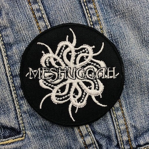 Op de afbeelding: Een ronde, zwarte patch met het woord "MESHUGGAH" geborduurd in het wit, samen met een wit, gestileerd ontwerp. De patch is bevestigd op een spijkerjasje.