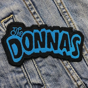 Puede incluir: Parche bordado rectangular con el texto "The Donnas" en azul, contorneado en negro. El parche está sobre un fondo de tela vaquera.