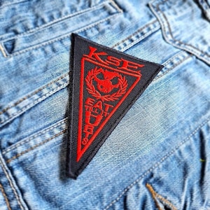May include: A black triangular patch with red embroidered text and a bull graphic, reading "KSE EAT IT UP". The patch is sewn onto a light blue denim jacket, showcasing the texture of the fabric and the patch's design.