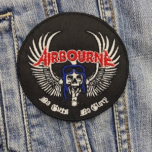 Puede incluir: Un parche redondo negro con la palabra "AIRBOURNE" en letras rojas, alas blancas y una calavera con pelo azul. El texto "No Guts No Glory" está debajo de la calavera en blanco. El parche está sobre una chaqueta vaquera.