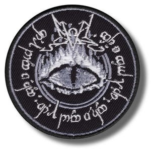 May include: Round black patch featuring a detailed white and gray design. The central image is an eye with flames and script around the edge. The patch is likely an embroidered or printed item, suitable for clothing or accessories.