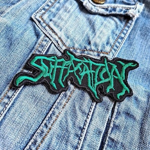 Può includere: Una toppa ricamata nera e verde con la scritta "STARVATION" in un carattere stilizzato. La toppa è cucita su una giacca di jeans blu chiaro. Il testo è verde con un contorno nero.