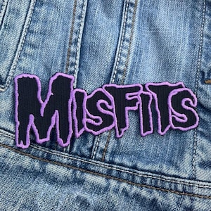 Pode incluir: Um patch preto com a palavra "MISFITS" em uma fonte roxa estilizada. O patch é costurado em uma jaqueta jeans azul. As letras têm um efeito de gotejamento, dando-lhe uma estética punk rock.