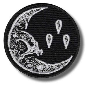 May include: A round black patch with a white embroidered crescent moon design and three white teardrop shapes. The moon has detailed, textured embroidery. The patch has a black border and is likely an iron-on or sew-on patch.