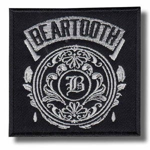 May include: A square, black patch featuring the word "BEARTOOTH" in white, arched lettering. Beneath, a circular design with an ornate, white floral pattern surrounds a white letter "B". The patch has a white border and is likely for clothing.