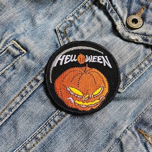 May include: A circular black patch featuring the word "HELLOWEEN" in white above an orange pumpkin with yellow features. The patch is sewn onto a blue denim jacket.
