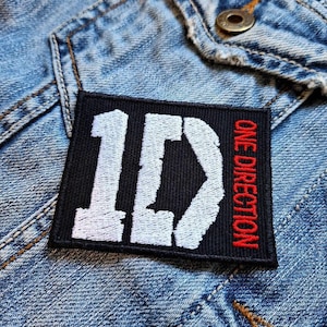 May include: A black rectangular patch featuring the white letters "1D" and the red text "ONE DIRECTION". The patch is attached to a light blue denim jacket.