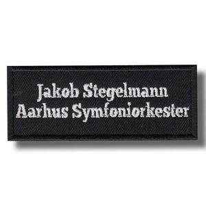 Op de afbeelding: Een zwart rechthoekig embleem met wit geborduurde tekst. De tekst luidt "Jakob Stiegelmann Aarhus Symfoniorkester". De patch is waarschijnlijk een naamplaatje of uniformidentificatie.