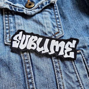 Op de afbeelding: Een zwart-wit geborduurde patch met het woord "SUBLIME" in een gestileerd lettertype. De patch is rechthoekig en is geplaatst op een blauw spijkerjasje.