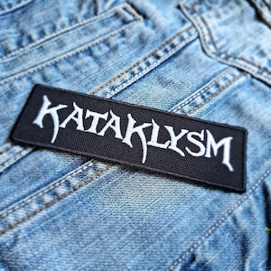 May include: A black rectangular patch with the word "KATAKLYSM" embroidered in white, stylized lettering. The patch is sewn onto a light blue denim fabric, with visible stitching and seams. The patch is likely for a jacket or bag.