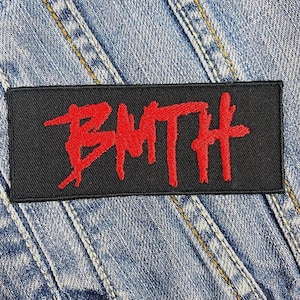 Puede incluir: Un parche rectangular negro con las letras rojas bordadas "BMTH". El parche está cosido sobre una chaqueta vaquera azul claro. Las letras están en una fuente estilizada, manuscrita.