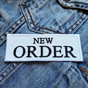 Puede incluir: Un parche rectangular blanco con las palabras "NEW ORDER" bordadas en negro, sobre un fondo de chaqueta vaquera. El parche tiene un diseño limpio y minimalista, con un enfoque en el texto en negrita.