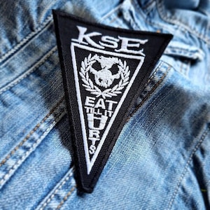 May include: Black and white triangular patch with the text "KSE" at the top and "EAT TILL IT DIES" below a skull and laurel wreath design. The patch is on a denim jacket.