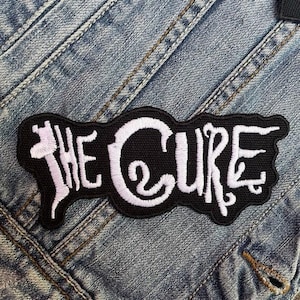 May include: A black embroidered patch featuring the band name "THE CURE" in white, decorative lettering. The patch is sewn onto a denim jacket. The patch has a distinctive, flowing shape.