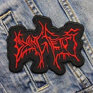 Op de afbeelding: Zwarte geborduurde patch met rode, gestileerde tekst. De patch heeft een gekartelde omtrek en is op een spijkerjasje genaaid. De rode tekst lijkt een bandlogo of ontwerp te zijn.