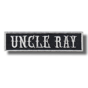 Pode incluir: Patch retangular preto com o texto bordado "UNCLE RAY" em branco, com letras góticas. O patch tem uma borda branca e um efeito de sombra. O patch é provavelmente para roupas ou acessórios.
