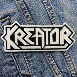 Op de afbeelding: Zwart-wit geborduurde patch met het woord "KREATOR" in witte, vette letters. De patch heeft een witte rand en is geplaatst op een blauwe denim achtergrond.