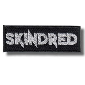 May include: A rectangular black embroidered patch with the word "SKINDRED" in white, stylized lettering. The patch has a textured appearance and a subtle shadow.
