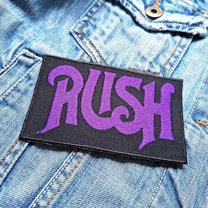 Pode incluir: Um patch retangular preto com a palavra "RUSH" bordada em roxo, com uma fonte estilizada. O patch está colocado sobre uma jaqueta jeans azul clara, com costuras e um bolso visíveis.