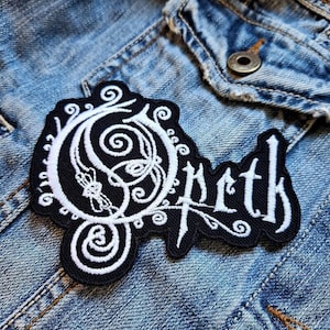 May include: Black and white embroidered patch with the word "Opeth" in a gothic font. The patch features an ornate design with swirls and a stylized letter "O". The patch is placed on a denim jacket.