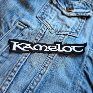 May include: A black and white embroidered patch with the word "Kamelot" in a stylized font. The patch is rectangular with curved edges and is placed on a denim jacket.