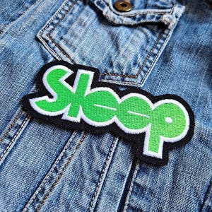 May include: A rectangular patch with the word "Sleep" in green, outlined in white, on a black background. The patch is sewn onto a denim jacket, with visible stitching and a pocket.
