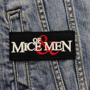 Op de afbeelding: Zwart rechthoekig embleem met de tekst "MICE MEN" in het wit, met "OF" erboven en een rode ampersand. De patch is op een spijkerjas genaaid.