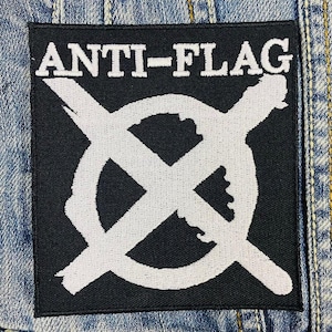 Może przedstawiać: Kwadratowa czarna naszywka z białym napisem "ANTI-FLAG" u góry. Biały symbol, okrąg z X, znajduje się na środku. Naszywka jest na tle dżinsu.
