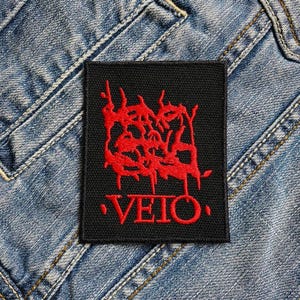 Op de afbeelding: Zwart vierkantige patch met een rood abstract ontwerp en het woord "VEIO" geborduurd in rood. De patch wordt getoond op een blauwe denim achtergrond, die de textuur van de stof benadrukt.