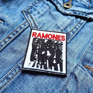 May include: A rectangular patch with the word "RAMONES" in red block letters above a black and white graphic of a band. The patch is sewn onto a blue denim jacket.