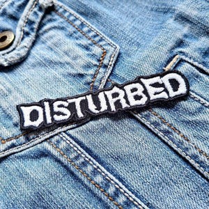 May include: A rectangular embroidered patch in black and white, featuring the word "DISTURBED" in a bold, stylized font. The patch is sewn onto a light blue denim jacket, visible in the background.
