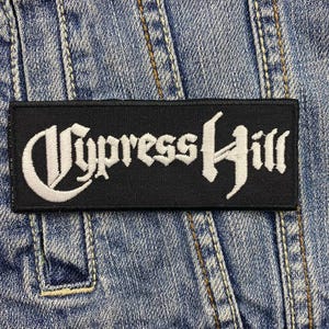 Op de afbeelding: Een rechthoekige zwarte patch met de witte tekst "Cypress Hill" in een gotisch lettertype. De patch is genaaid op een blauw spijkerjasje, met zichtbare stiksels en naden.