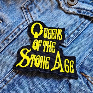 Może przedstawiać: Czarna i żółta naszywka z napisem "Queens of the Stone Age" w stylizowanej czcionce. Naszywka jest przyszyta do dżinsowej kurtki.